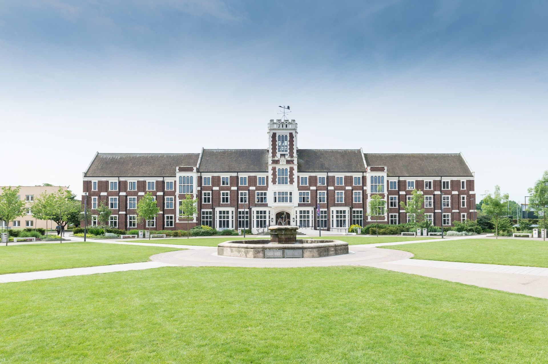 Large brick building with a central tower, fountain, and green lawn.