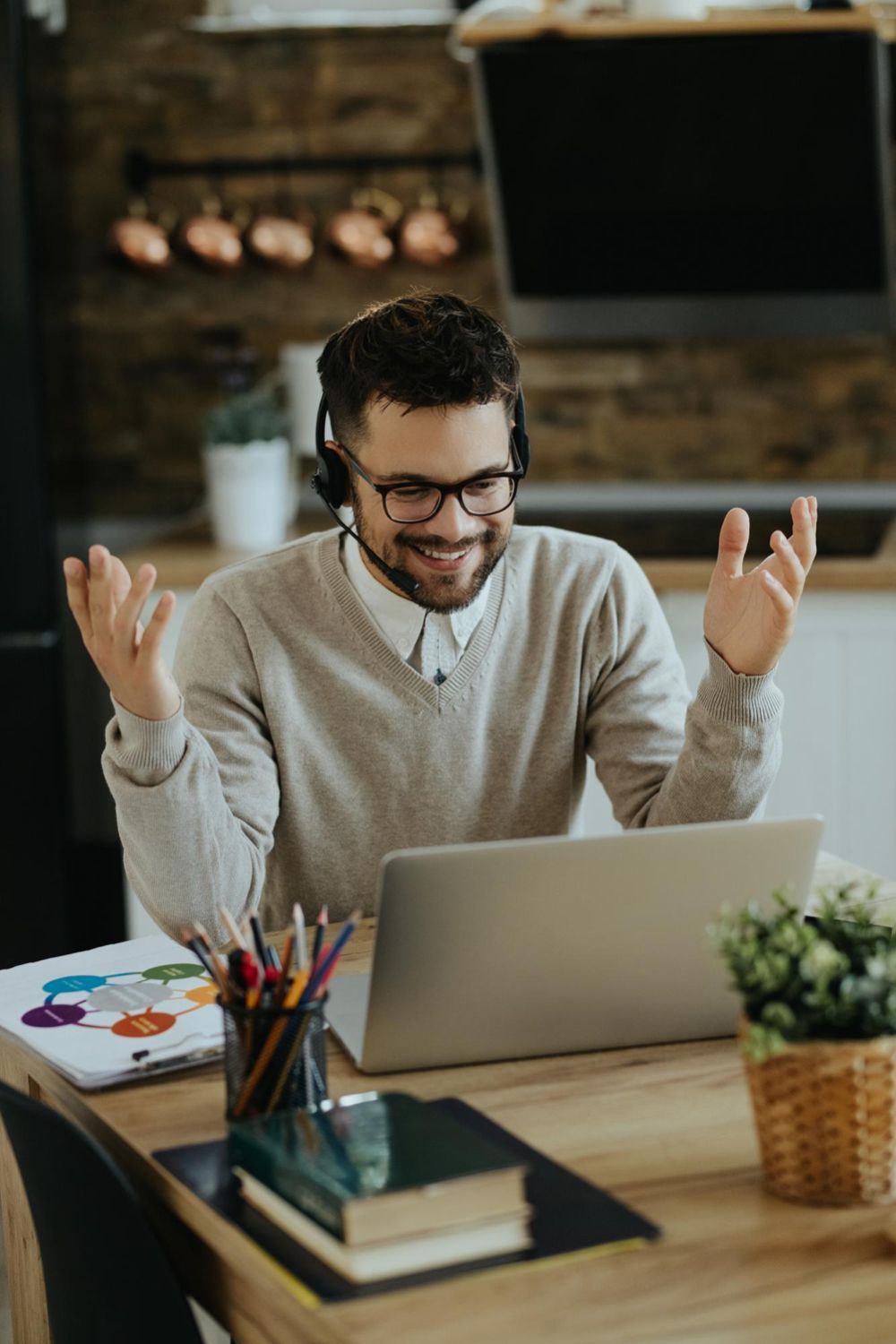 Man with glasses and headset smiles while video conferencing on a laptop at a kitchen table.