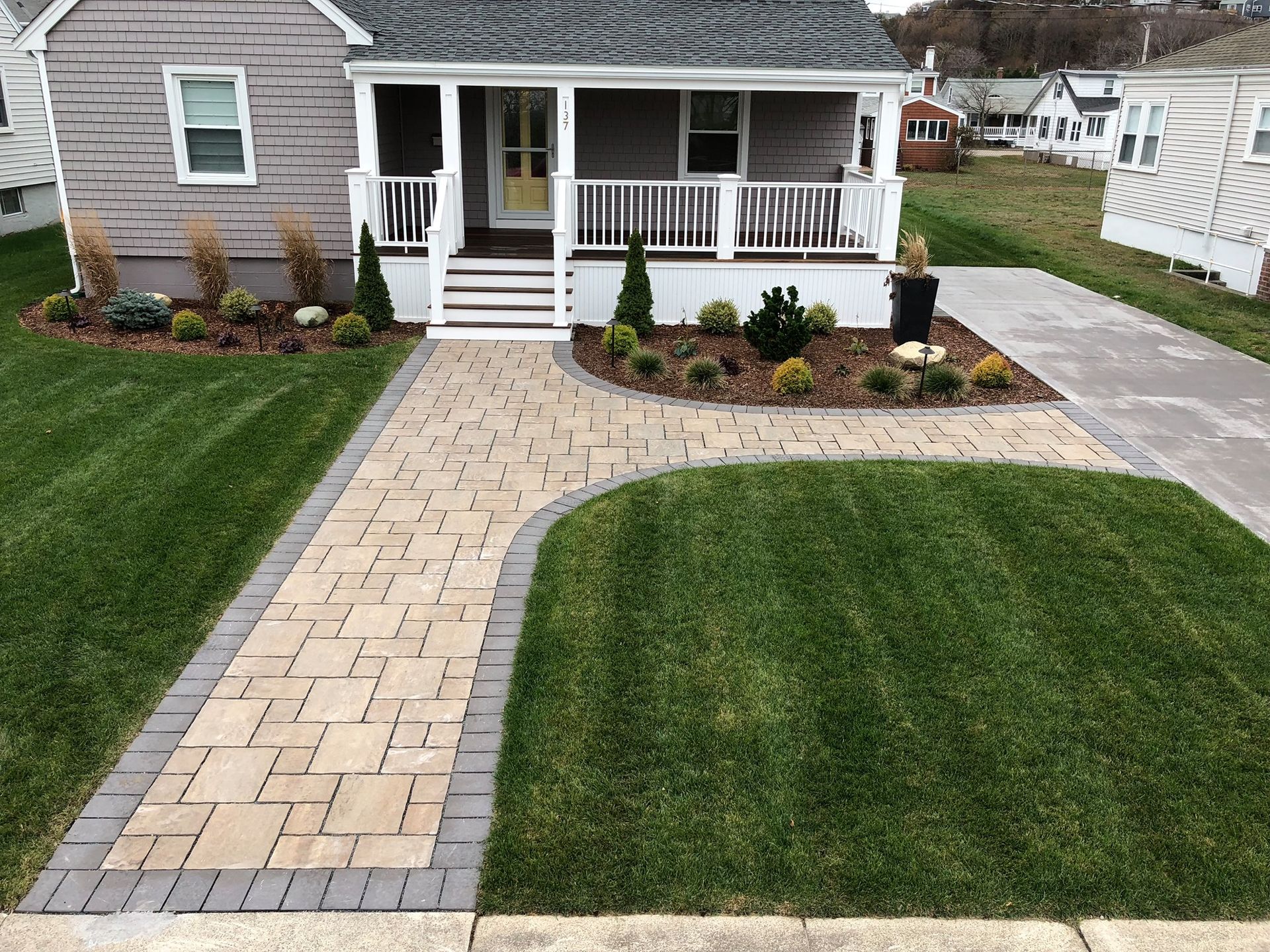 A brick walkway leading to a house with a porch. The yard is very well maintained.