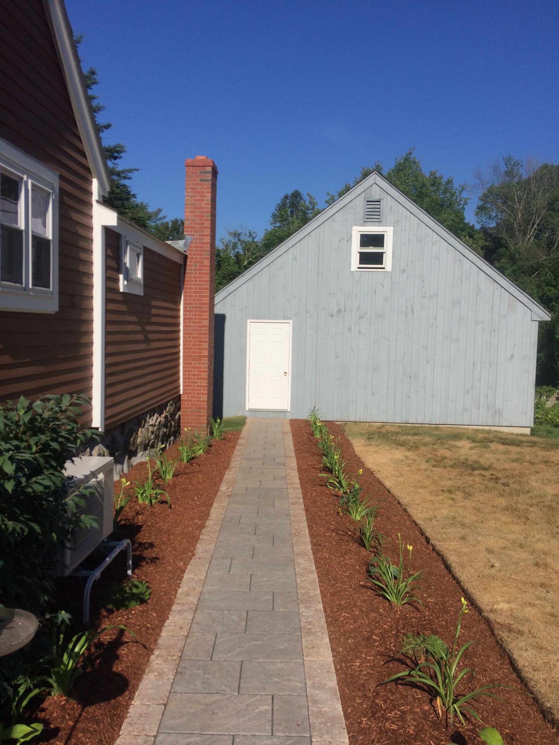 Brick path leads from a red house, past flower beds to a light blue shed on a sunny day.