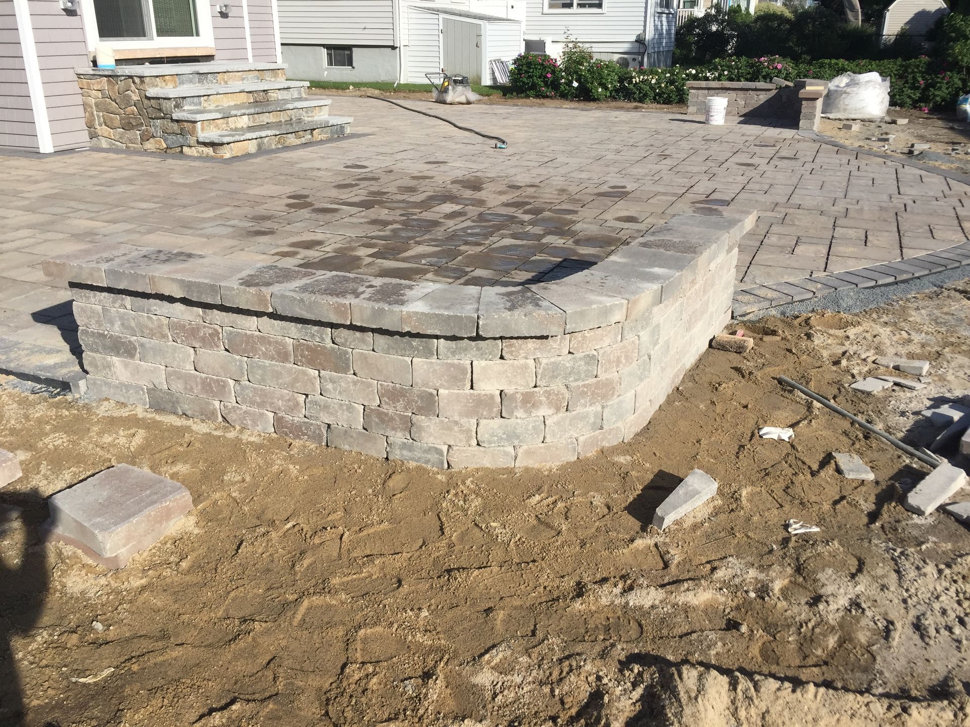 Curved brick retaining wall built on a paved patio, surrounded by dirt, bricks, and a house.