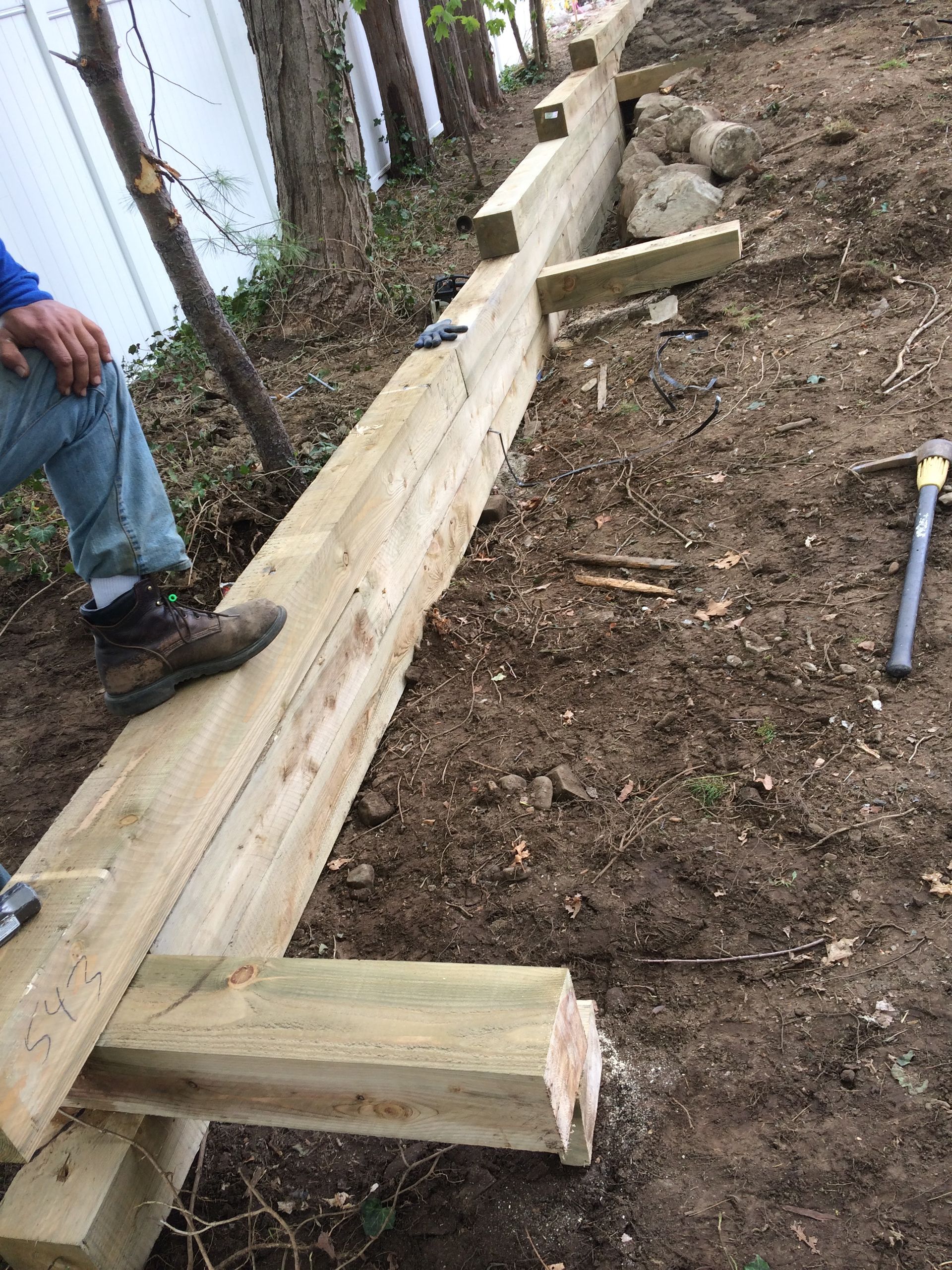 Man standing on wooden beam, building a retaining wall outdoors.