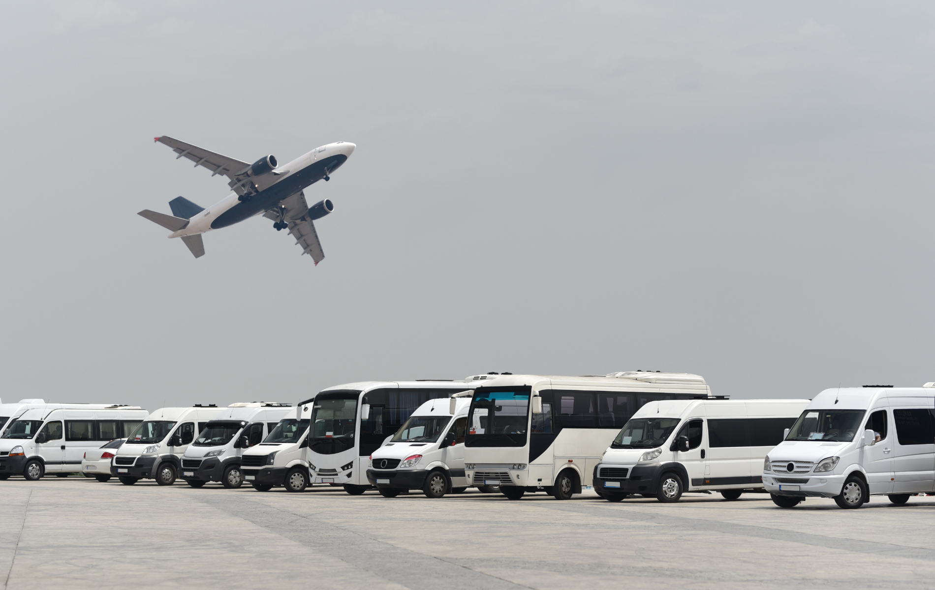 A plane is flying over a row of buses and vans.