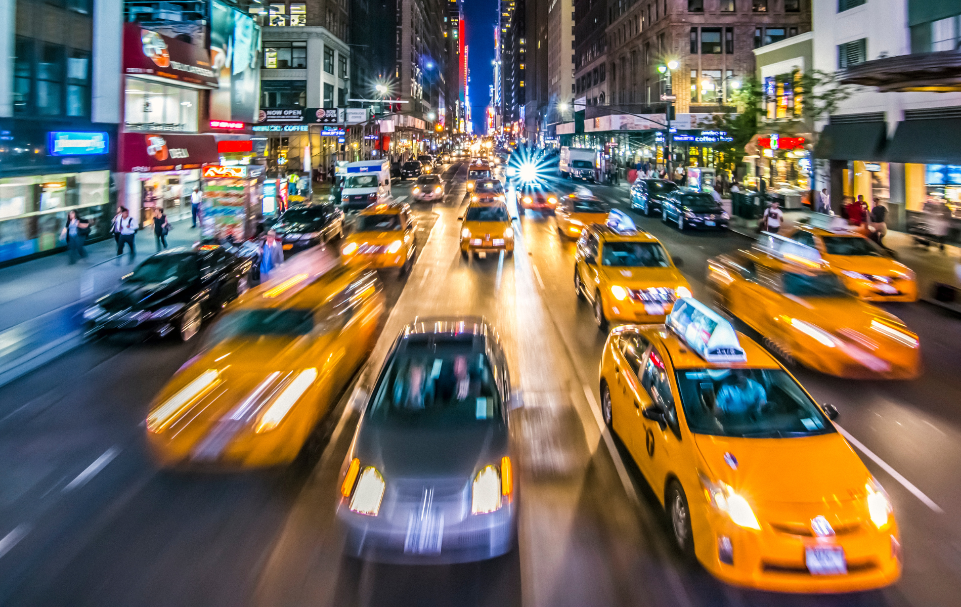 A bunch of yellow taxis are driving down a city street at night.