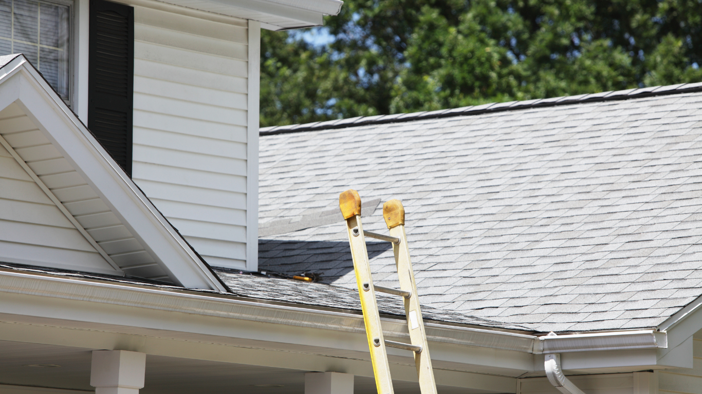 Two yellow ladders lean against a gray house roof near a white eave.