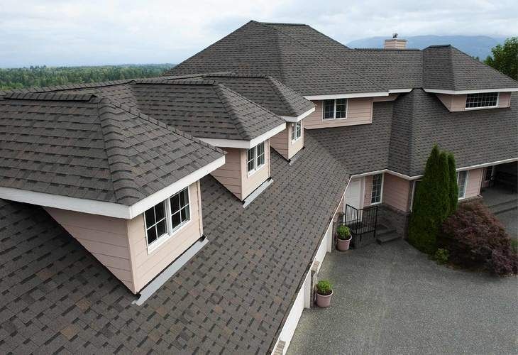Aerial view of a large gray-roofed suburban house with a gravel driveway and distant mountains.