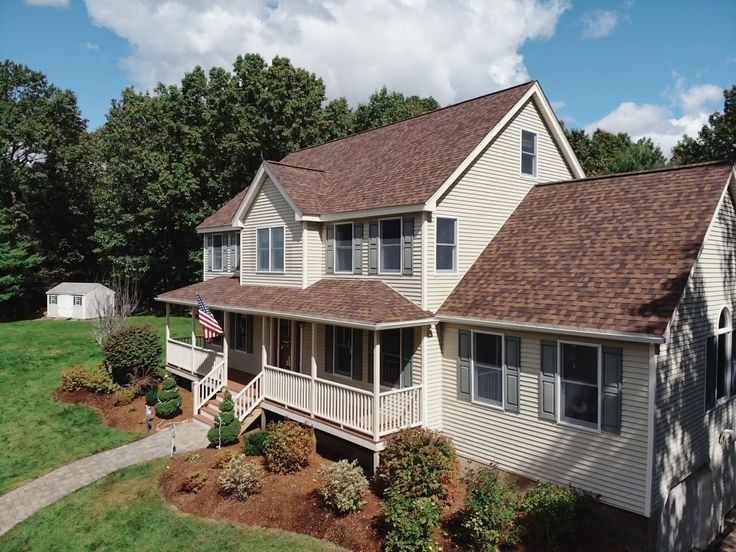Two-story beige house with red roof, white porch, and landscaped yard on a sunny day