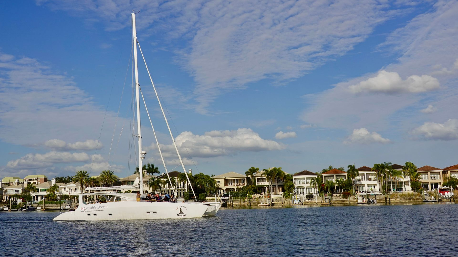 White sailboat on calm blue water with waterfront homes and palm trees under a partly cloudy sky