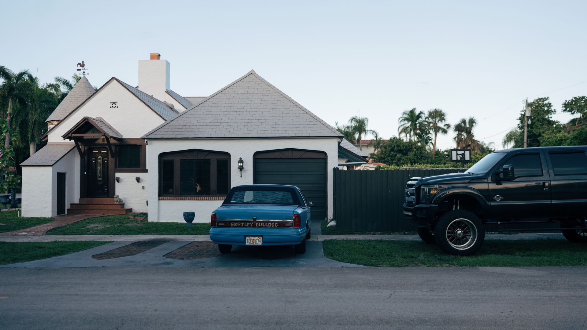 Suburban house with two parked cars, one blue in the driveway and one black pickup at the curb.