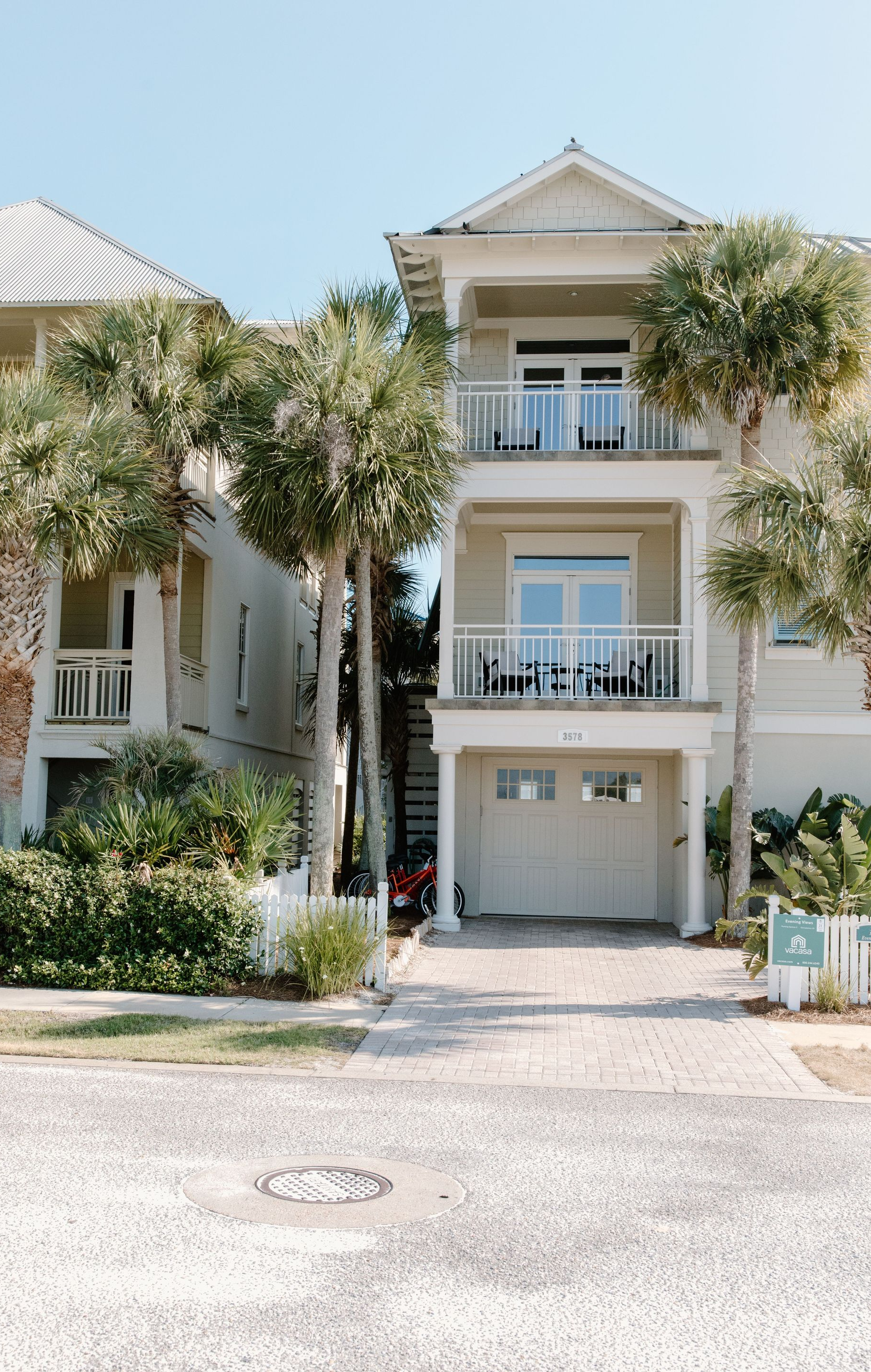 Two-story white townhouse with balconies, garage, and lush trees along a paved street