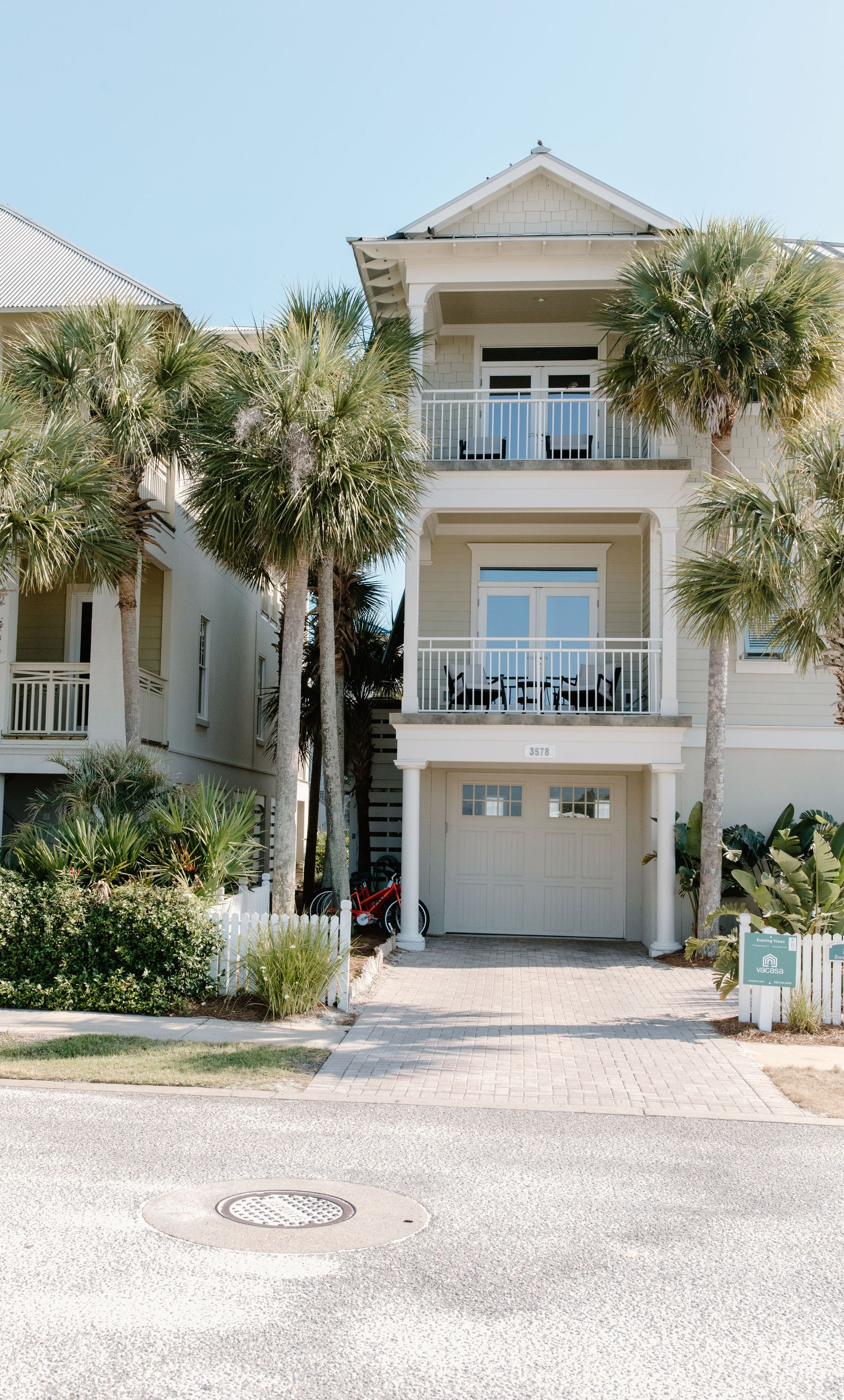 Two-story white townhouse with balconies, garage, and lush trees along a paved street
