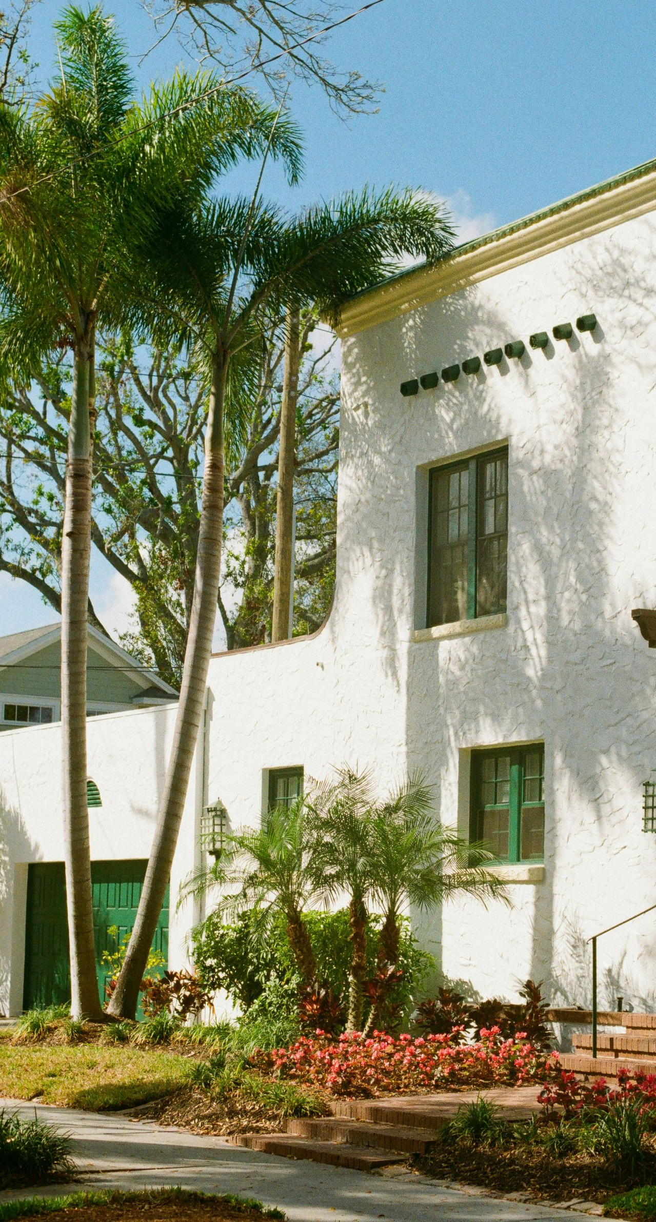 White building with green-trimmed windows and tall palm trees beside a landscaped garden