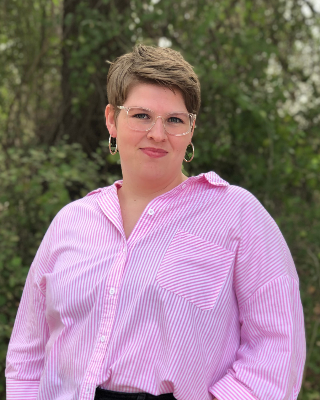 A woman wearing glasses and a pink striped shirt is standing in front of trees.