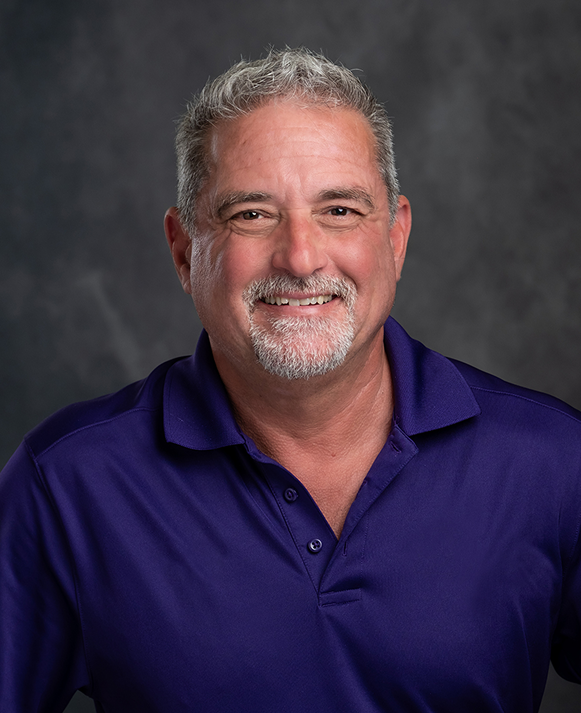 A man with a beard is wearing a purple shirt and smiling.