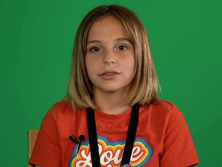 A young girl wearing a red t-shirt and suspenders is sitting in front of a green screen.