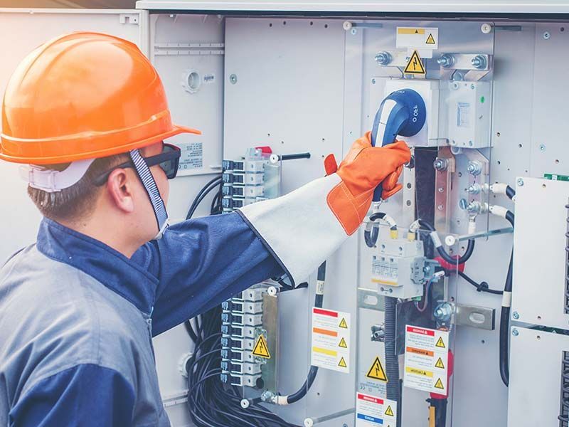 A man wearing a hard hat and gloves is working on an electrical box.