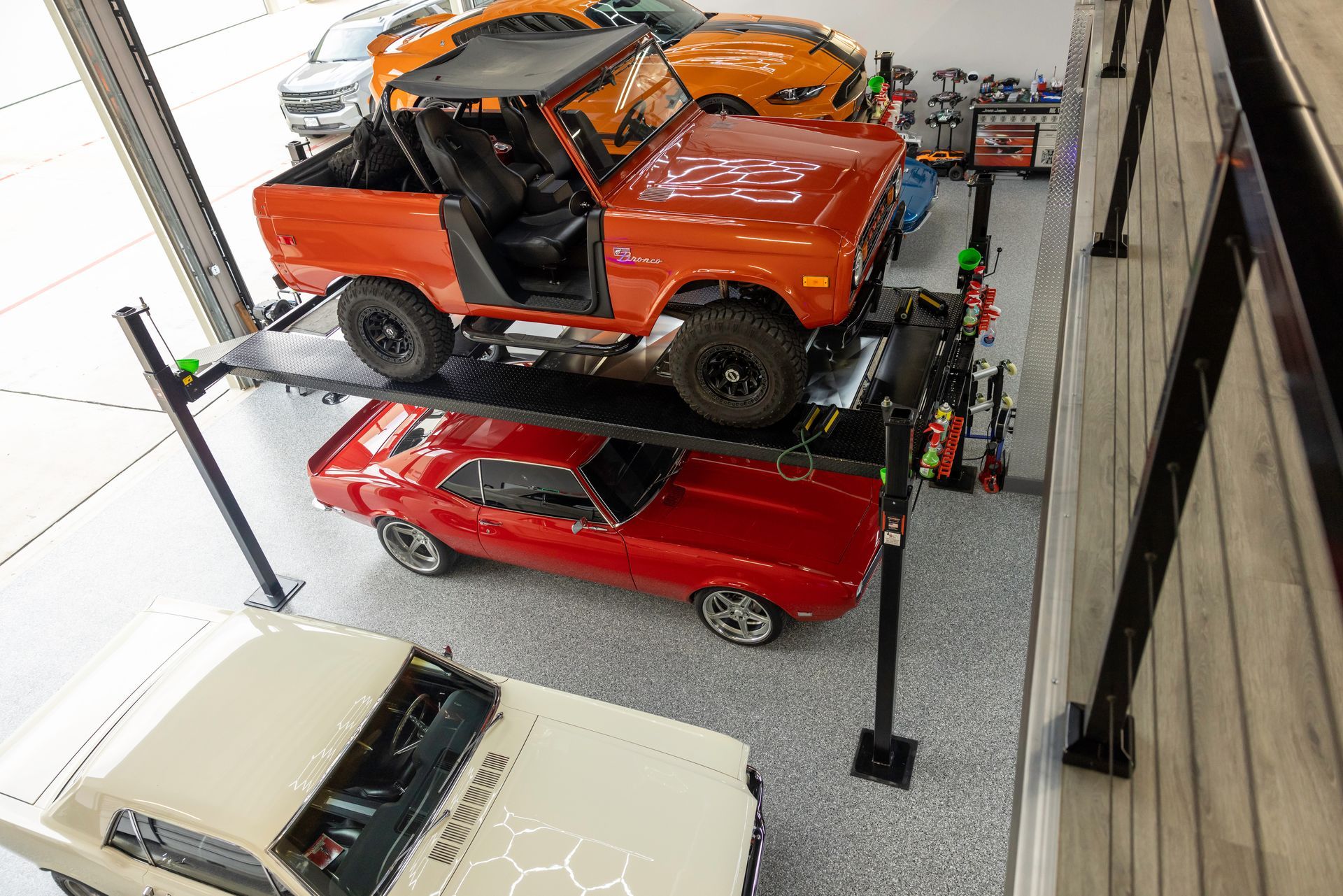 Orange Bronco parked above a red muscle car in a garage with other vehicles. | Auto Motive