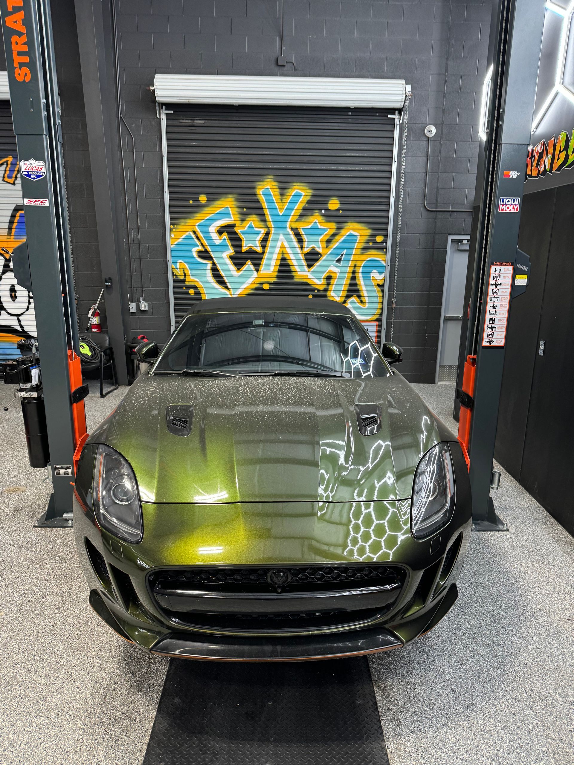Green Jaguar sports car in a garage with graffiti and a closed garage door. | Auto Motive