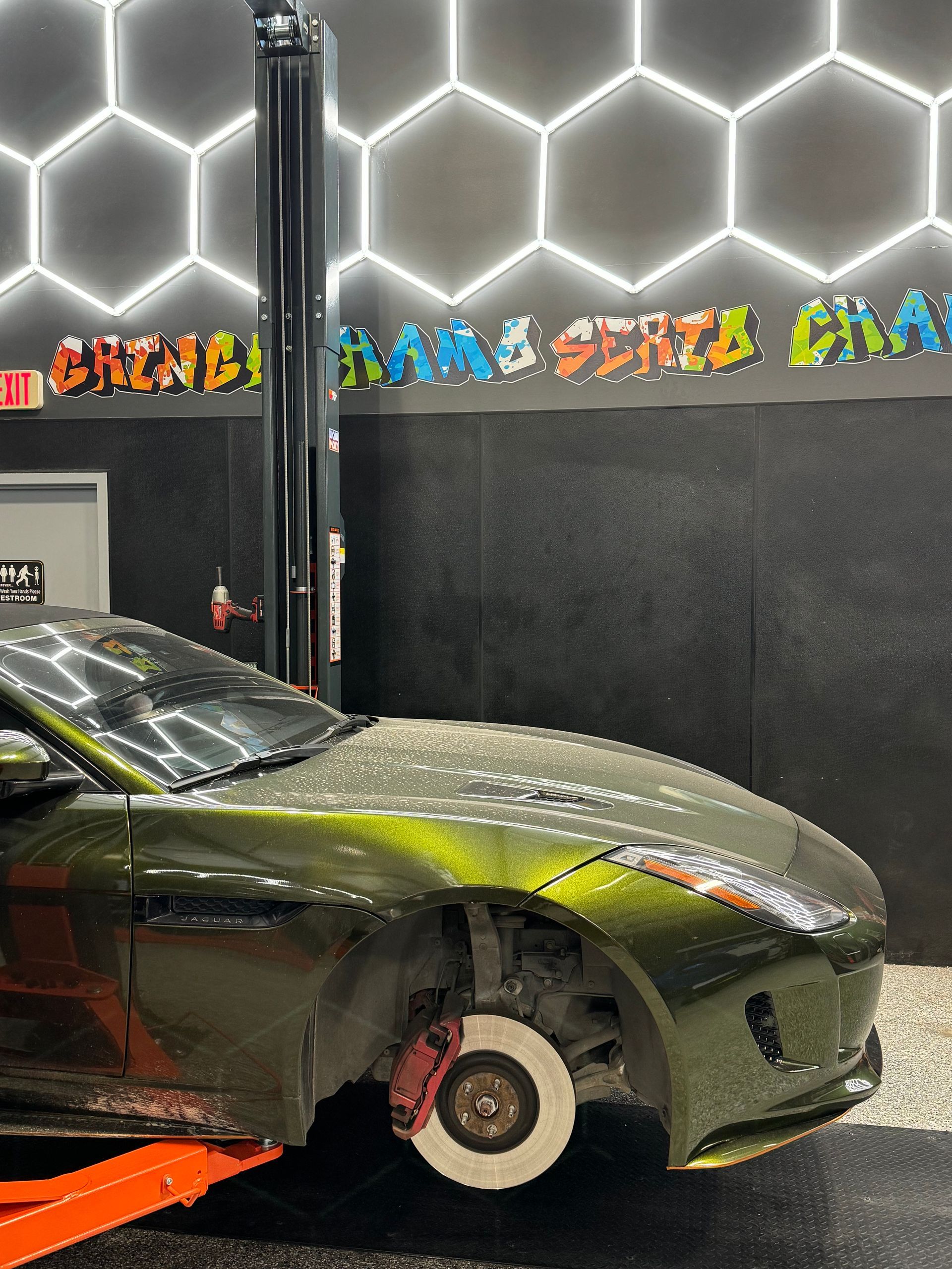 Green sports car on a lift in a garage; front tire removed, red brake caliper visible. Hexagon lights on the wall. | Auto Motive