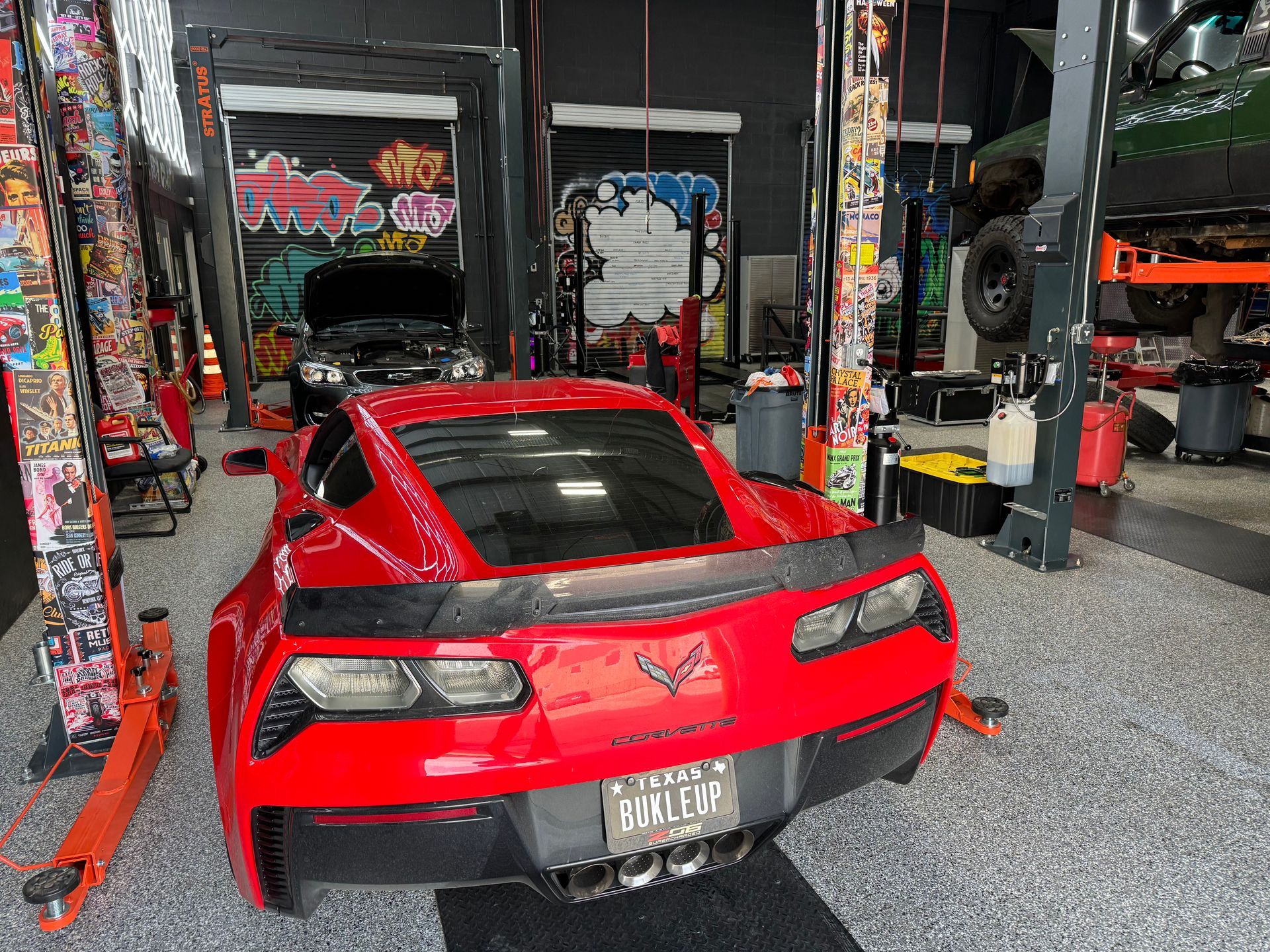 Red Corvette sports car in a garage, next to a lift, with tools and a vehicle in the background. | Auto Motive