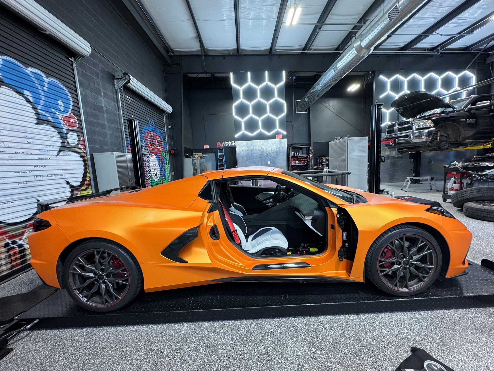 Orange sports car with open door in garage, gray walls, and honeycomb lights. | Auto Motive
