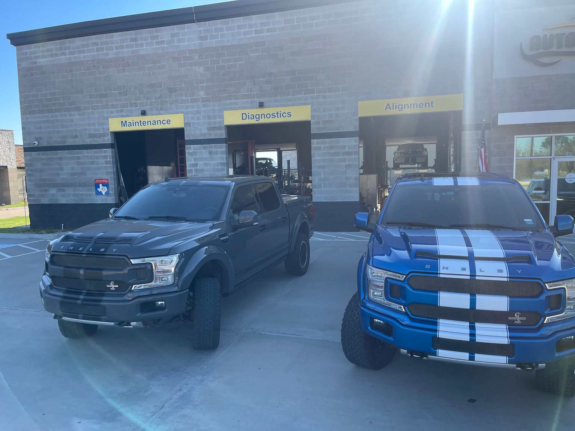 Two Ford trucks parked outside a building: one dark gray, one blue with white stripes. Sunny day. | Auto Motive