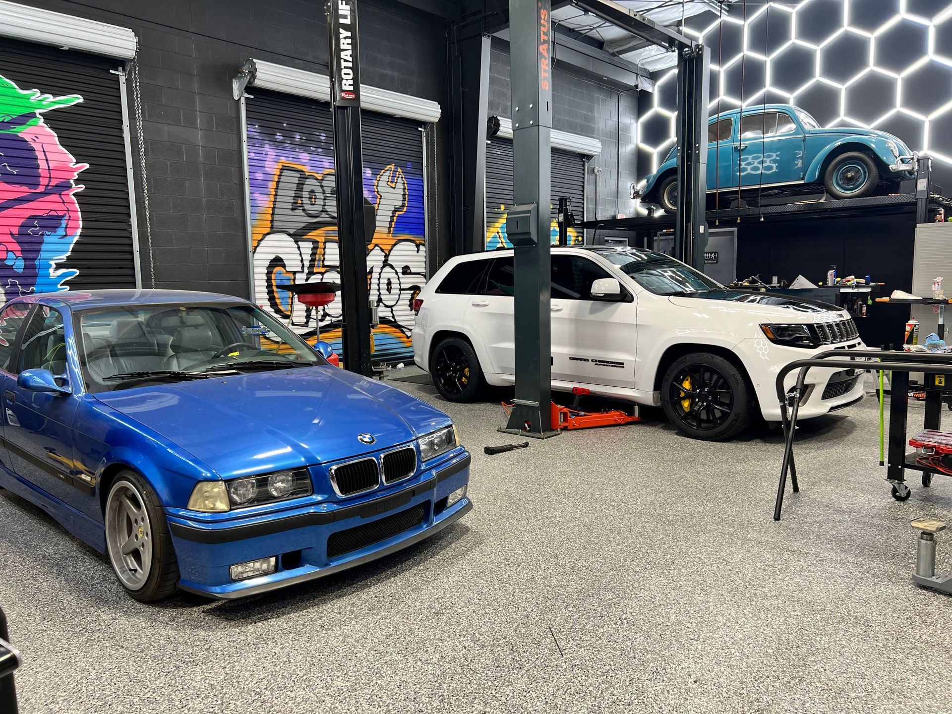 A blue BMW, white Jeep, and blue vintage car in a garage with graffiti and neon lights. | Auto Motive
