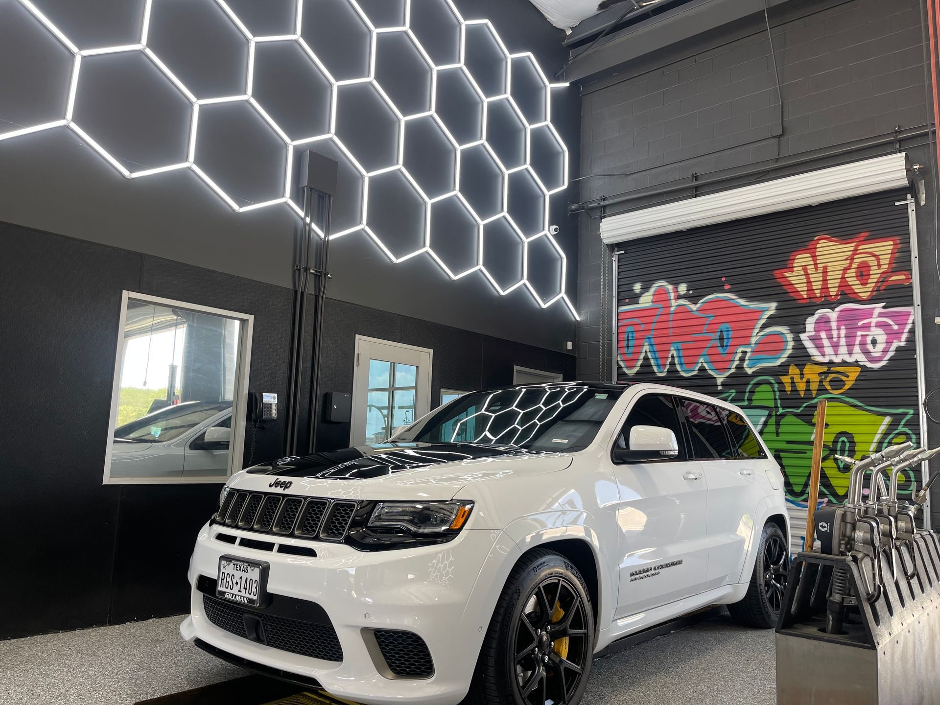 White SUV parked inside a garage with honeycomb-shaped lights on the wall and graffiti art on the door. | Auto Motive