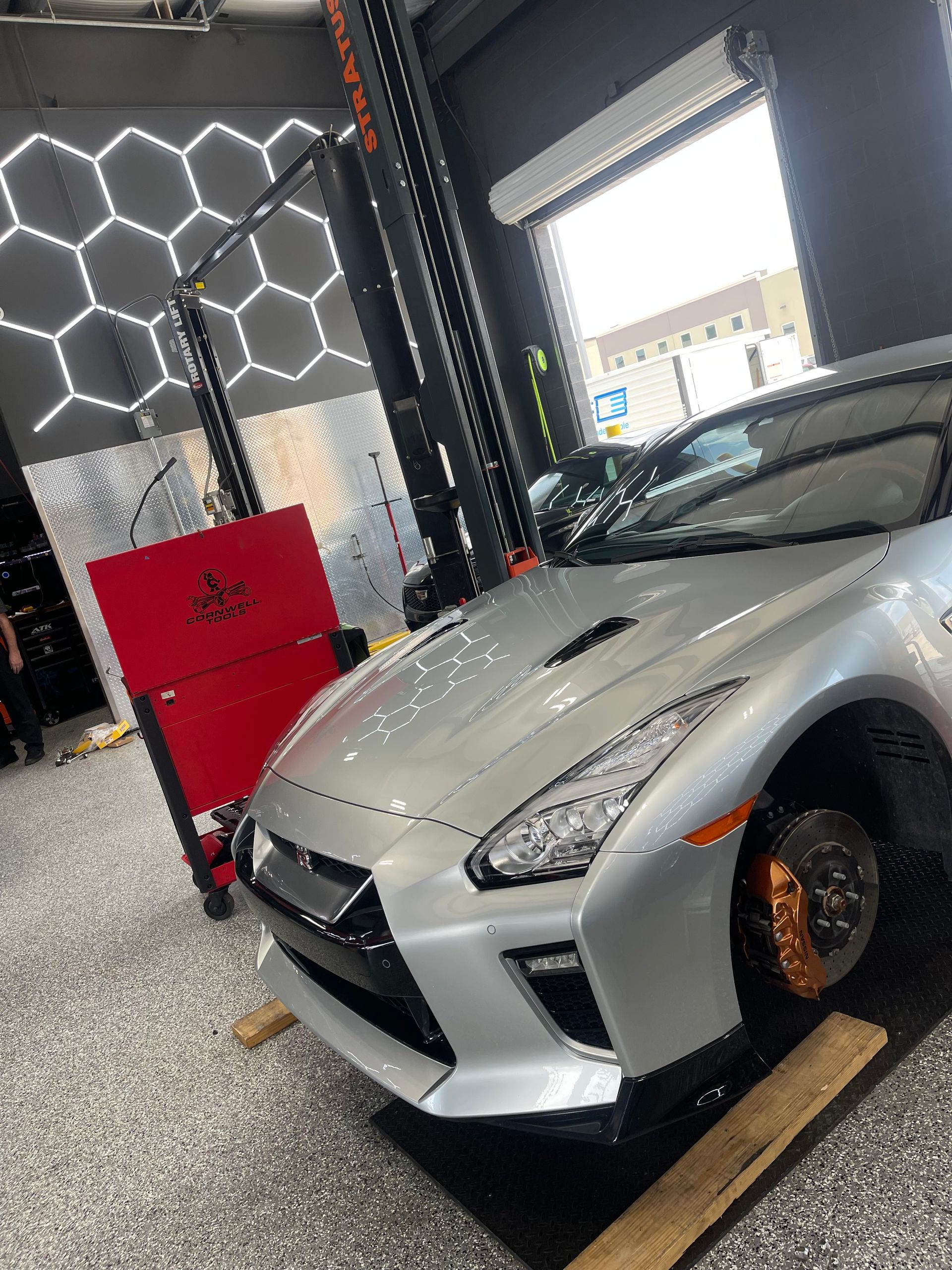 Silver Nissan GT-R in a garage, brake calipers visible, on a lift. Hexagon lights and red tool cabinet in background. | Auto Motive