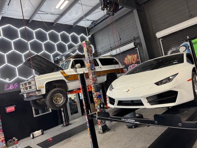 A white Lamborghini and a raised truck inside a modern auto shop. Hexagon lights on the wall.