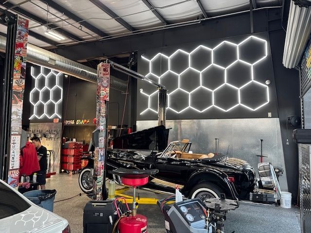 An auto repair shop with a car on a lift. White honeycomb-shaped lights decorate the black wall.