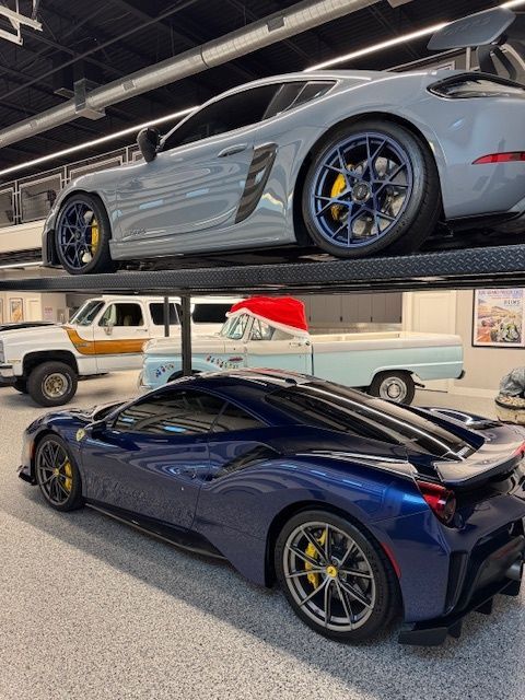 Blue Ferrari and gray Porsche in a garage with classic trucks on an upper level.