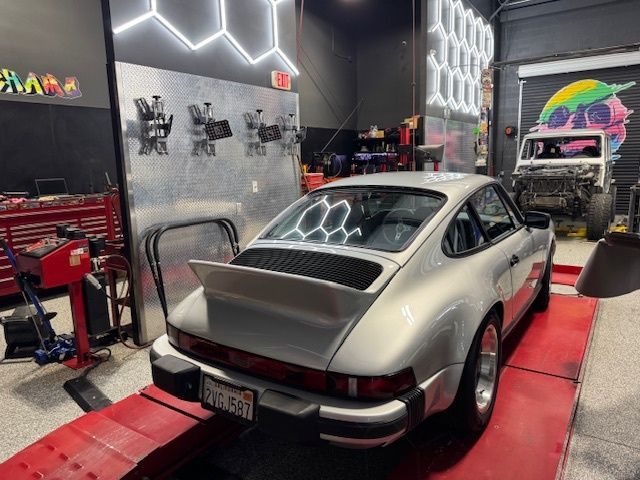 Silver Porsche 911 on a lift inside a garage, with honeycomb lights, tools, and another car in the background.