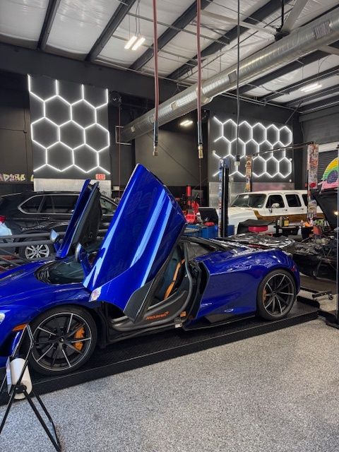 Blue sports car with doors open in a garage, orange interior, black wheels, next to honeycomb lights.