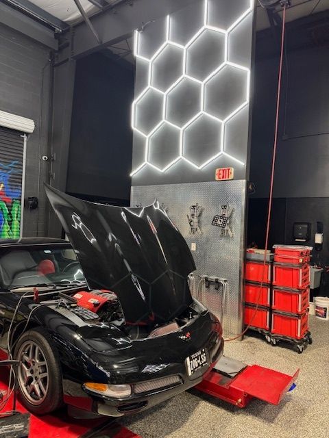 Black Corvette on a lift with open hood, in a garage with hexagon-light wall.
