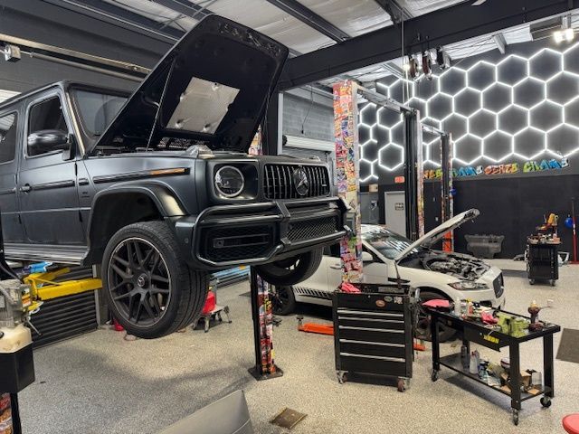 A matte black Mercedes G-Wagon raised on a lift in a garage, hood open, with another car in the background.