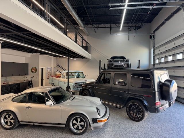 Silver Porsche, black Mercedes SUV, vintage truck, and black car on a lift in a modern garage.