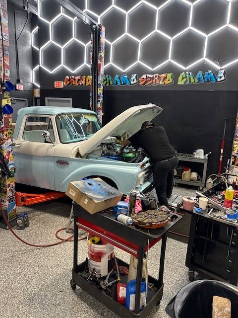 Mechanic working on a light blue classic pickup truck inside a garage. Tools and parts cart in foreground.