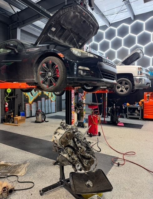 Black car on a lift in an auto shop with its engine on the floor. A white truck is also in the bay.