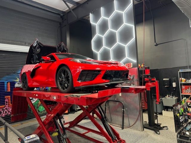 Red Corvette on a red lift inside a garage. Honeycomb neon lights on the wall.