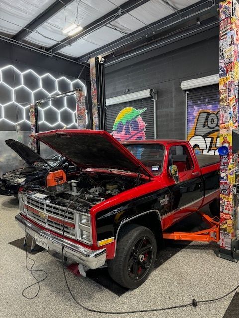 Red and black pickup truck raised on a lift in a garage, hood open for repair.