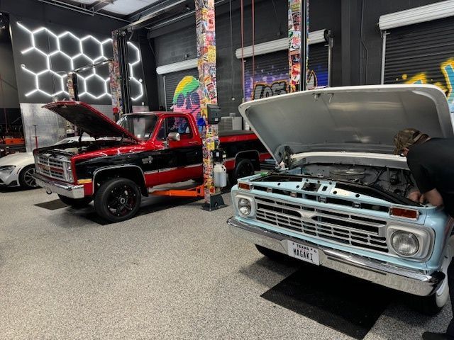 Two classic trucks with open hoods in a garage, being worked on.
