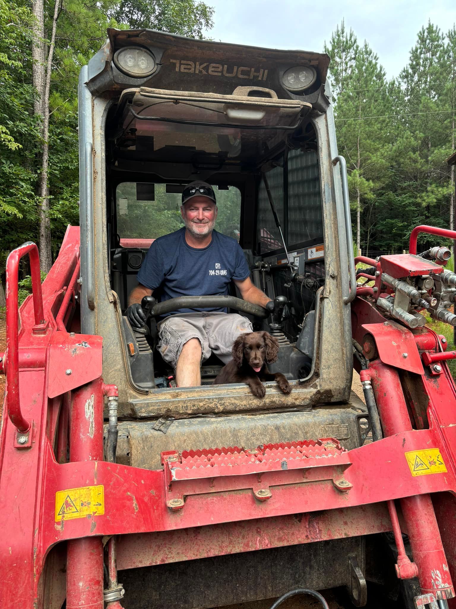 Man in a red tractor cab with a dog. Both look at the camera. Outdoors, woods in the background.