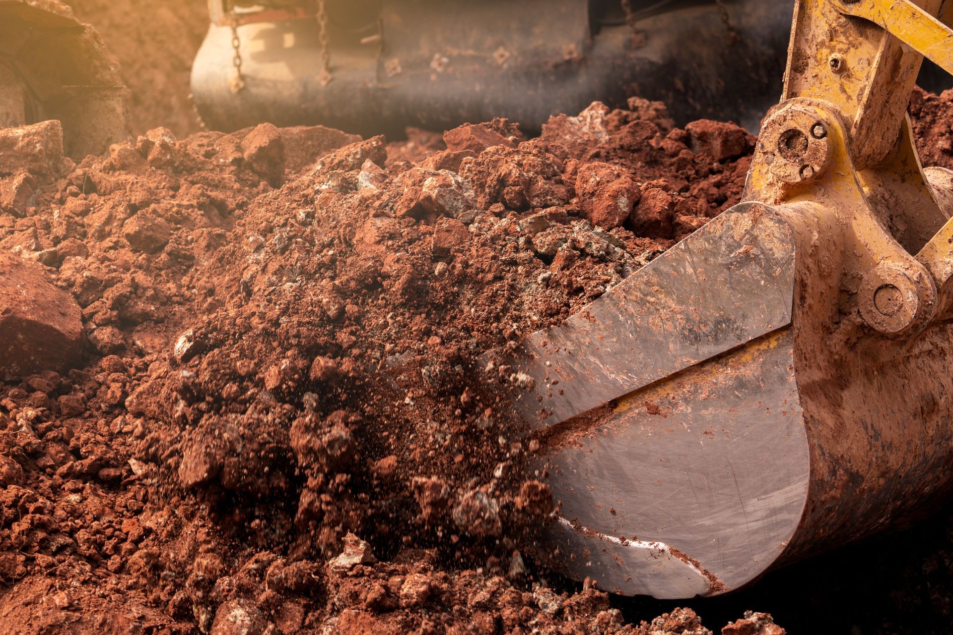 Excavator scoop filled with red-brown soil, likely construction site.