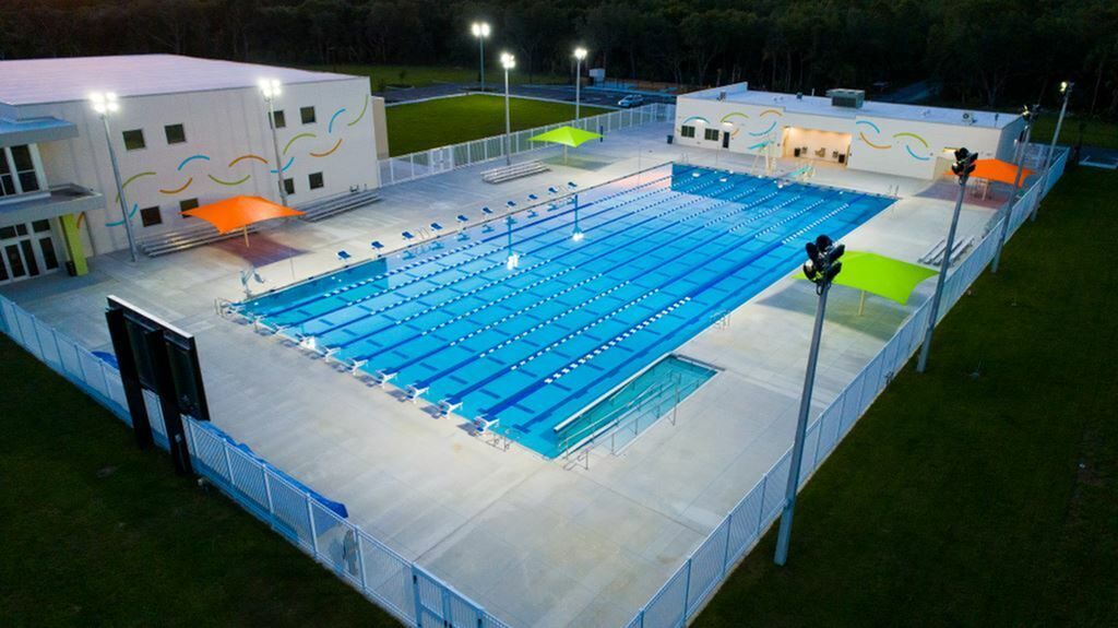 Aerial view of a lit outdoor swimming pool with lanes, surrounded by buildings and grass at night