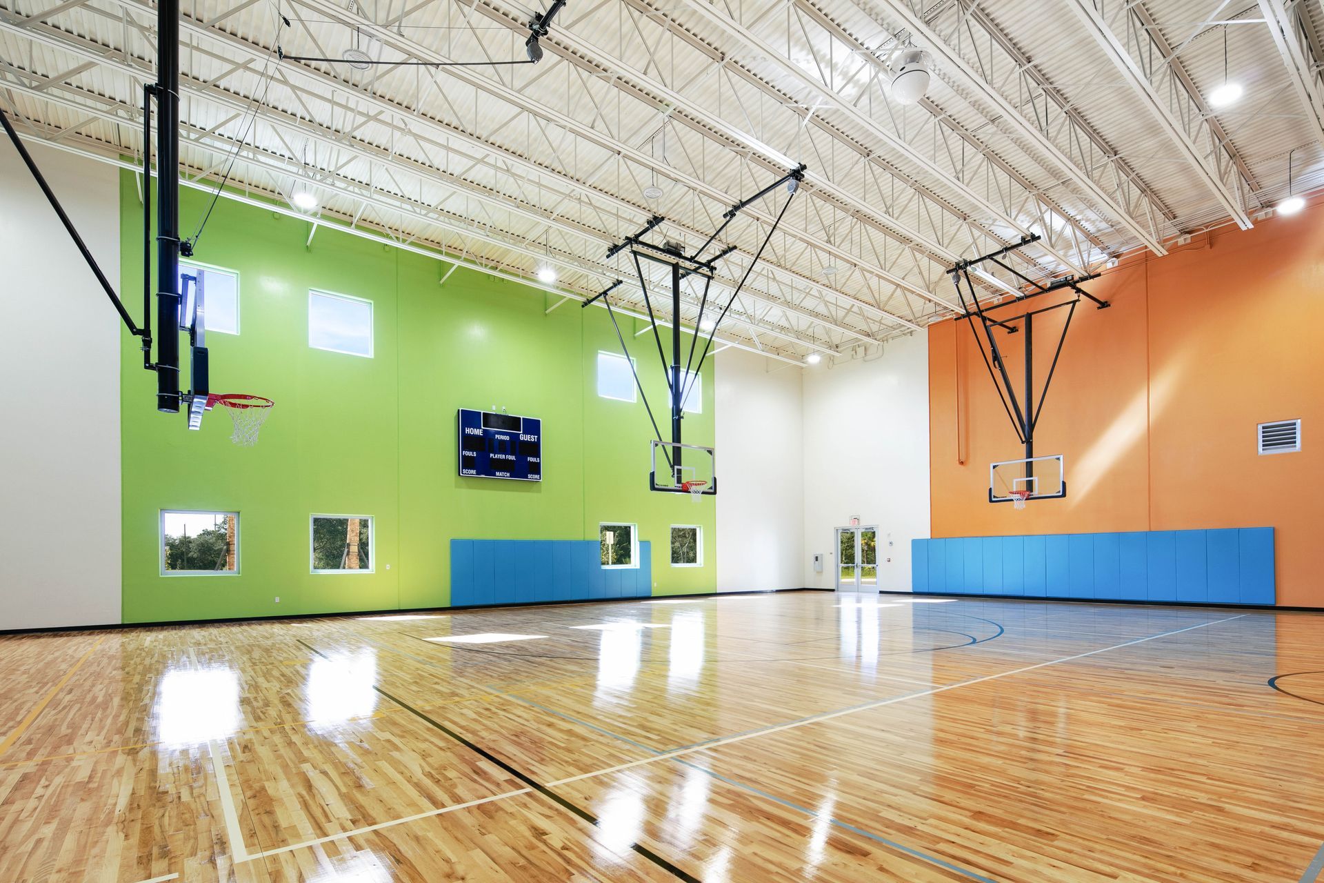 Empty indoor basketball gym with polished wooden floor, green wall, hoops, and bright ceiling lights
