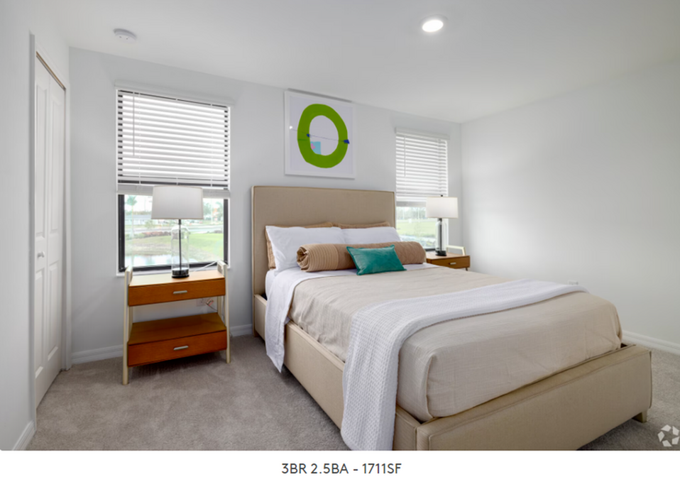 Bright bedroom with white walls, a beige bed, and two windows beside a small wooden nightstand.