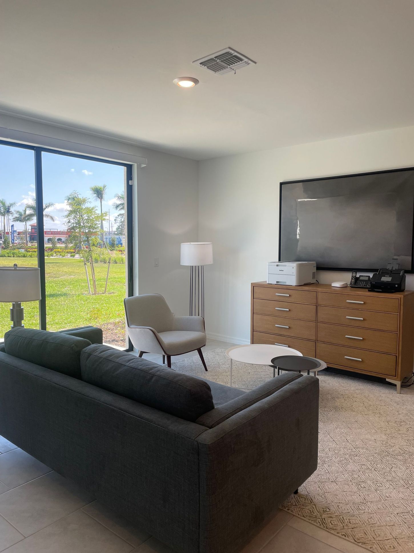 Modern living room with gray sofa, armchair, TV, dresser, and large window view of greenery