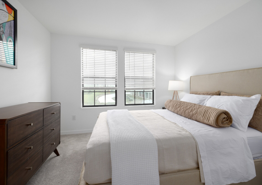 Bright bedroom with white bedding, beige headboard, dresser, and two windows with blinds