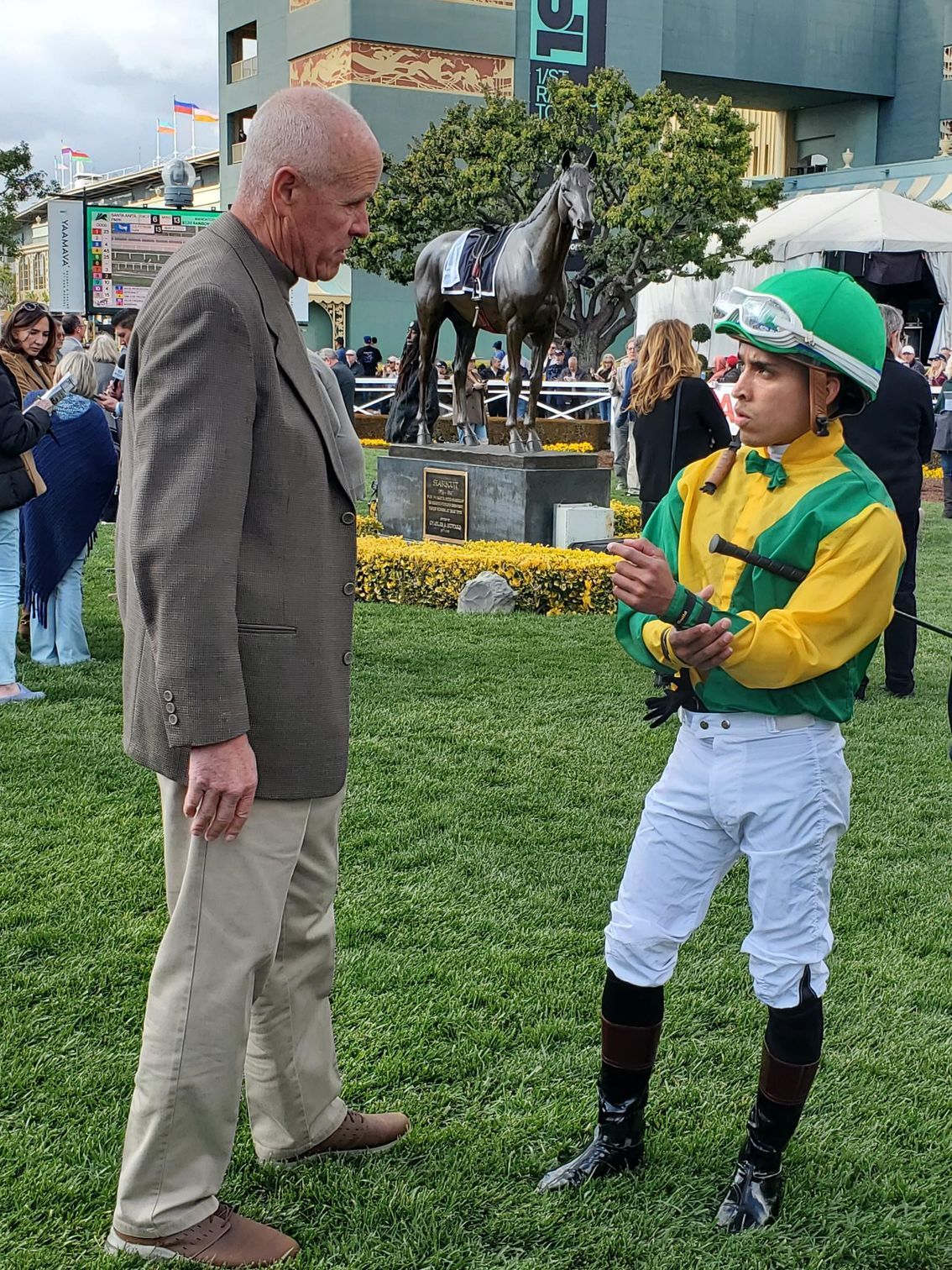 A man and a jockey are talking in a field with a statue of a horse in the background